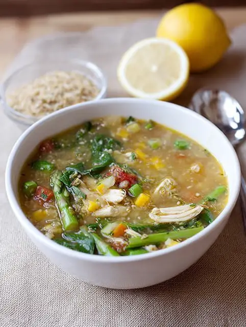 Bowl of chicken soup with spring vegetables, lemon wedges, and fresh basil on a wooden table.