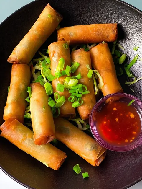 Crispy vegetable spring rolls served on a plate with dipping sauce and herbs.