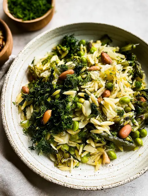 A vibrant bowl of spring orzo salad with asparagus, green olives, artichokes, and fresh herbs, topped with lemon zest.