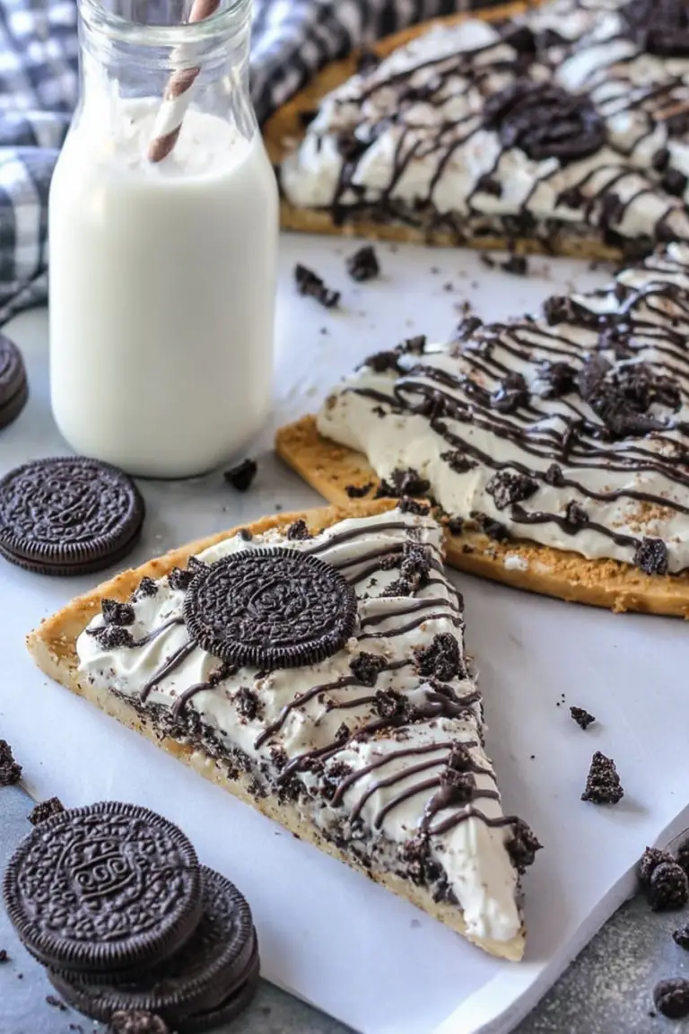 Close-up of cookies & cream dessert pizza topped with cream cheese icing, chopped Oreos, and white chocolate drizzle.
