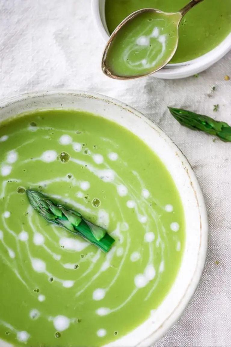 Creamy asparagus soup garnished with fresh tarragon and asparagus tips, served in a white bowl on a wooden table.