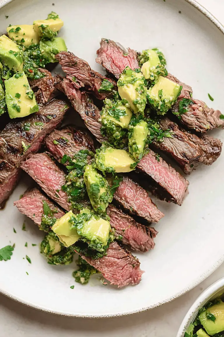 Grilled flank steak with avocado chimichurri, garnished with herbs and lime slices.