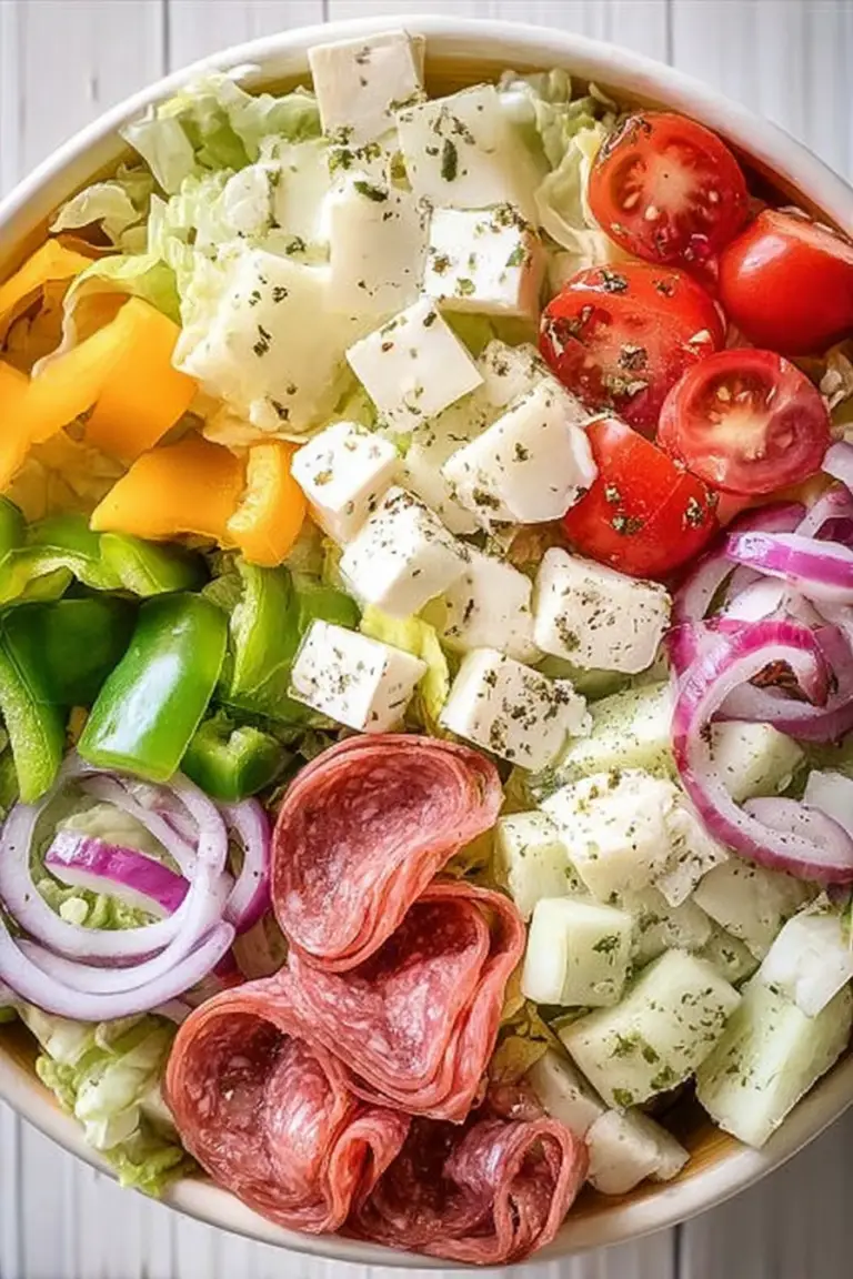 Italian sub salad with fresh ingredients including romaine lettuce, salami, provolone cheese, red peppers, and cucumbers in a large bowl.