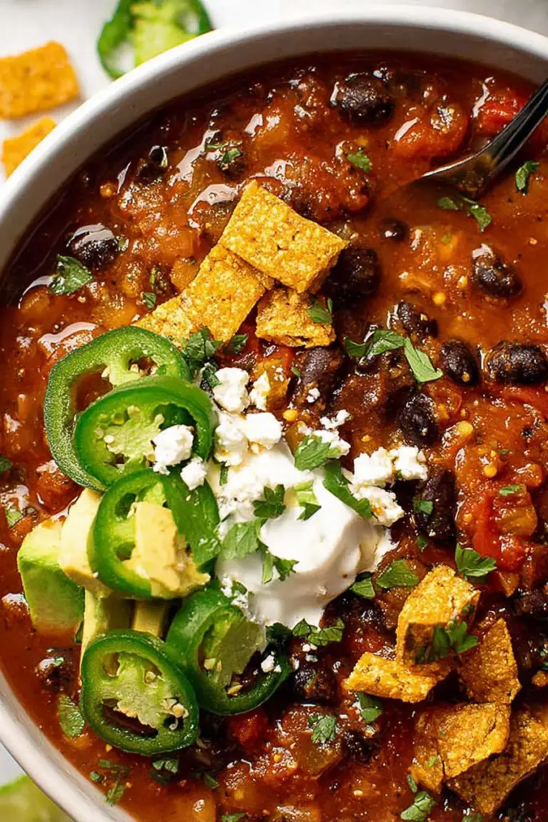 A delicious bowl of vegan black bean chili with garnishes of cilantro, jalapenos, and vegan sour cream, accompanied by Fritos.