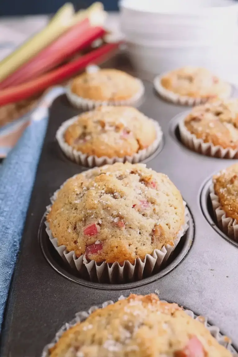 Freshly baked bakery style rhubarb muffins with coarse sugar on top and rhubarb stalks on a wooden table