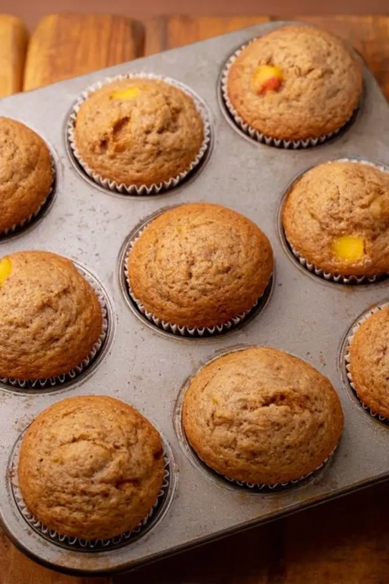 Freshly baked peach muffins with golden tops and peach chunks on a wooden table