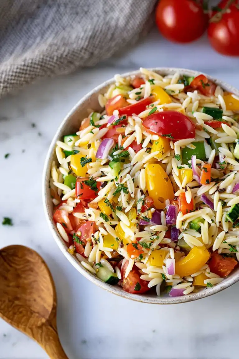 Colorful rainbow orzo salad served in a bowl on a picnic table with fresh vegetables around it.